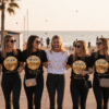A group of five women, all smiling and linking arms, walk toward the viewer on a wooden boardwalk near a beach at sunset. Four of the women wear matching black t-shirts with a smaller gold circular "TEAM Bride" logo. The woman in the center, the bride, wears a white t-shirt with a smaller gold "Bride to be" logo. All are wearing black jeans and sunglasses. Confetti falls around them.