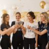 Lifestyle image of a group of happy women at a hen party, with the bride-to-be wearing a white short-sleeve 'Bride' t-shirt and the bridesmaids wearing matching navy blue 'Bridesmaid' t-shirts, promoting personalised hen party apparel and gifts.