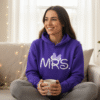 Lifestyle image of a woman sitting down and wearing a comfortable purple hoodie and trousers, clearly displaying the personalised white text "Mrs" on the front, ideal for bridal loungewear and personalised gifts.