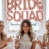 A celebratory hen party scene featuring three women sitting at a white table decorated with pink heart confetti and small gifts. The central woman wears a white 'Bride to be' t-shirt with gold script, while they all toast with glasses of champagne. Large rose gold 'BRIDE SQUAD' balloons are visible in the background against a bright window
