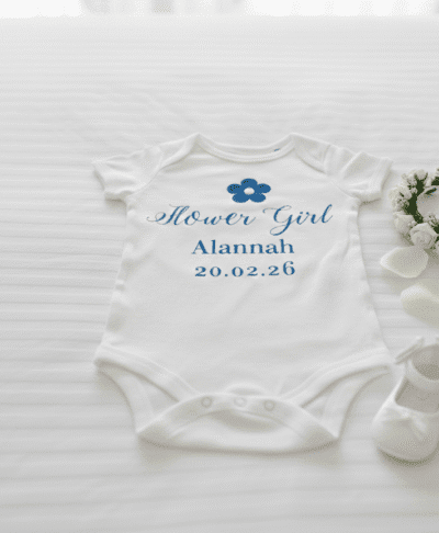 An overhead shot of a personalised white baby grow on a white bedspread, featuring turquoise text that reads 