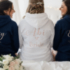 Bride in a white 'Mrs.' hoodie posing with her bridesmaids in matching 'Bridesmaid' hoodies, showing off wedding day preparation comfort wear.