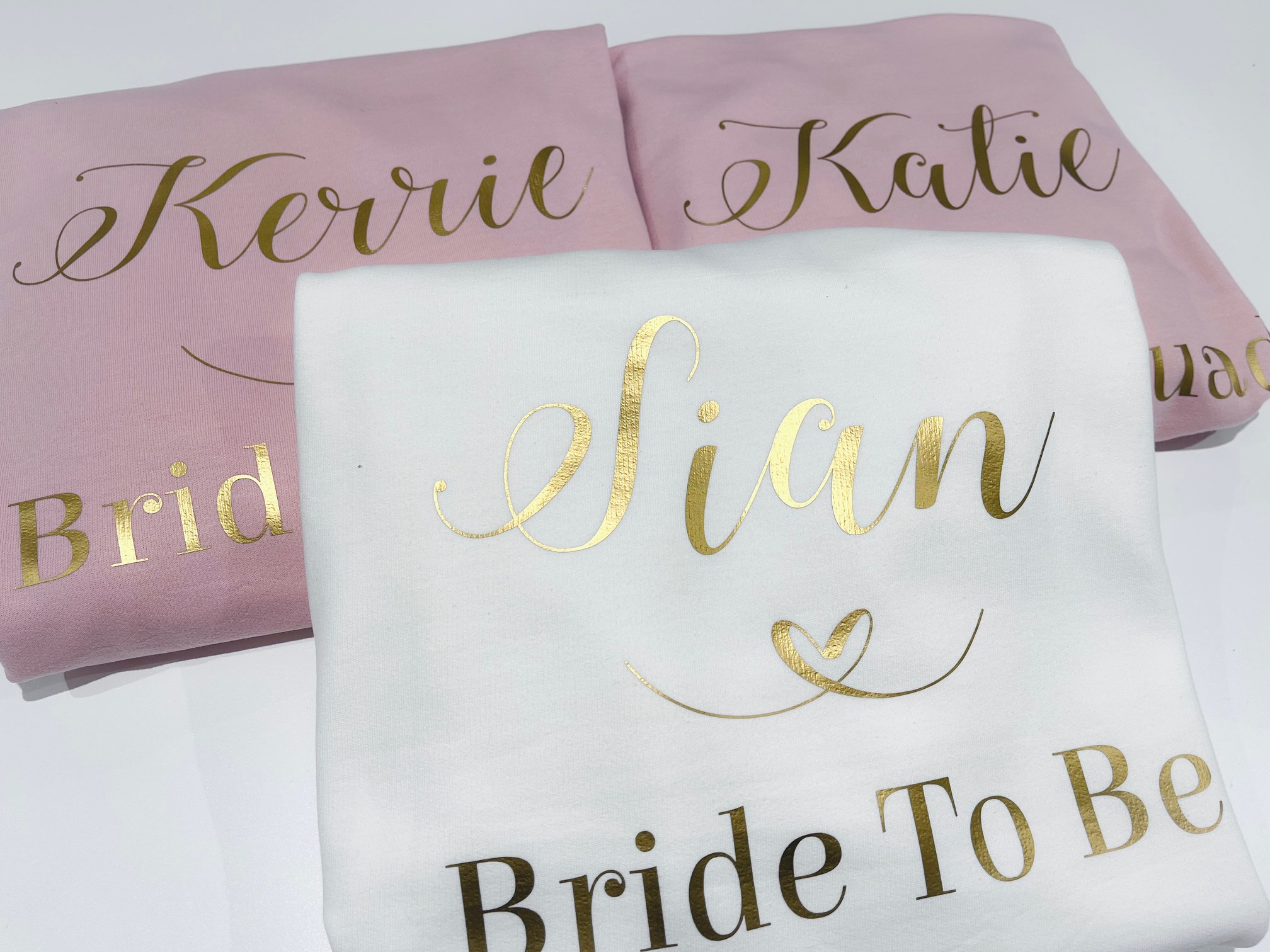 Close-up of three folded personalised hoodies: two light pink and one white. The white hoodie is in the foreground and features the name 'Sian' in large, metallic gold script, with 'Bride To Be' underneath. The pink hoodies in the background have names like 'Kerrie' and 'Katie' printed in gold script, with 'Bridesmaid' underneath.