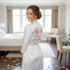 A young woman, presumably a bride, is standing slightly forward, wearing a white satin robe. The robes appear to be plain, with no lace trim. The back of the robes feature personalised text in a decorative script font, likely indicating their roles (e.g., "Bride," "Bridesmaid"). They are standing indoors in a bright, relaxed setting, possibly a hotel suite or dressing room.