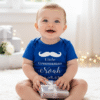 A joyful baby is sitting cross legged on a plush white rug, looking directly at the camera with a wide, happy smile. The baby is wearing a short-sleeved, bright blue bodysuit with white text and graphics that read: a mustache icon, "Little Groomsman Noah 30.06.23". The baby is holding a small, silver-wrapped gift box tied with a white ribbon in their hands. The background is softly blurred, showing a bright, airy space with white walls, soft lighting, and more silver-wrapped gifts and string lights, suggesting a festive or wedding related celebration.