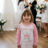 Lifestyle image of a flower girl smiling and getting ready in a bright room on the morning of a wedding, wearing white and pink personalised "Flower Girl" pyjamas, capturing a sweet moment of wedding preparation.