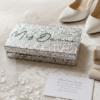 A silver glitter clutch bag is displayed on white, lace edged bridal fabric next to a pair of white pointed toe high heeled shoes, an open beige ring box containing a diamond engagement ring, and a small handwritten note on a white card.