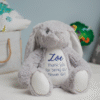 A plush grey personalised bunny sits centered on a white, fluffy rug. The bunny has long, floppy ears with white lining and a white patch on its belly and the soles of its feet. Printed in purple on its white belly is the name "Zoe" in a flowing script font. Below the name, the words "Thank you for being our Flower Girl" are stitched in a simple, clean font. The background is softly blurred, showing a blue wall.