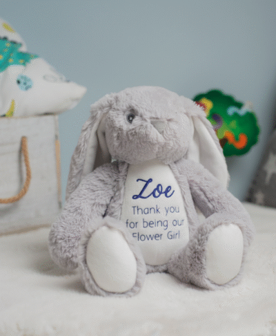A plush grey personalised bunny sits centered on a white, fluffy rug. The bunny has long, floppy ears with white lining and a white patch on its belly and the soles of its feet. Printed in purple on its white belly is the name "Zoe" in a flowing script font. Below the name, the words "Thank you for being our Flower Girl" are stitched in a simple, clean font. The background is softly blurred, showing a blue wall.