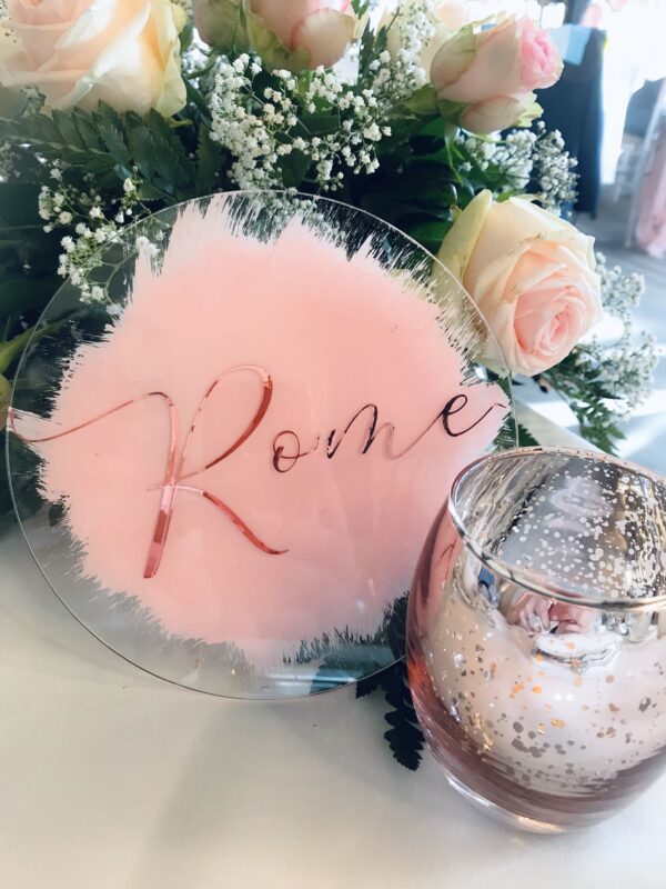 Clear acrylic circle table sign with pink painted background and rose gold lettering, styled with flowers and candles for wedding table décor.