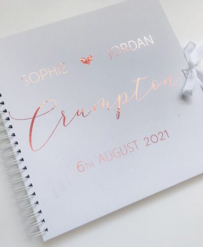 Personalised 12×12 wedding guest book with names, date, and surname in metallic script on a white cover with satin ribbon tie.