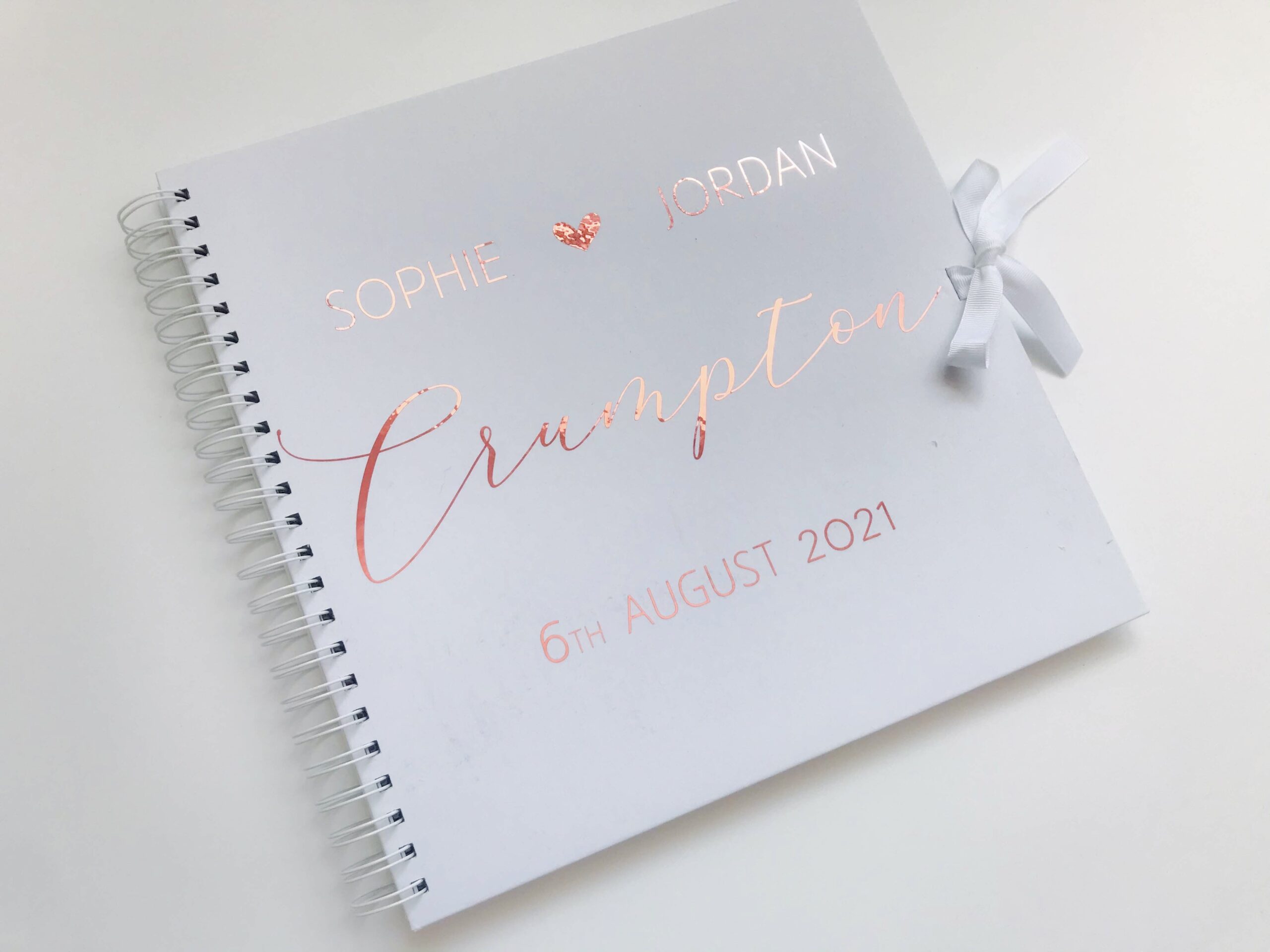 Personalised 12×12 wedding guest book with names, date, and surname in metallic script on a white cover with satin ribbon tie.
