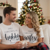 A happy husband and wife are posing together in a cosy Christmas setting. The wife is wearing short-sleeve white pajamas with red check trousers, and the top has navy text. The husband is wearing short-sleeve white pyjamas with grey check trousers, and his top has navy text. They are likely sitting near a decorated Christmas tree or fireplace, creating a festive, warm atmosphere.