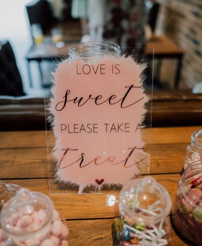 Clear acrylic sign reading “Love is Sweet Please Take a Treat” displayed on a wedding sweet table with jars of colourful sweets.