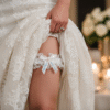 A close up view of a bride's leg shows a white lace garter with a satin ribbon bow and custom text placed high on her thigh. The garter's text is clearly readable in a light blue text.