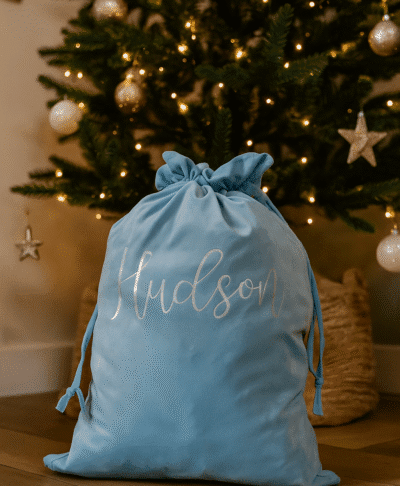 A light blue velvet Christmas sack with the name “Hudson” written in silver cursive sits on a wooden floor in front of a decorated Christmas tree adorned with gold and white ornaments and warm fairy lights.