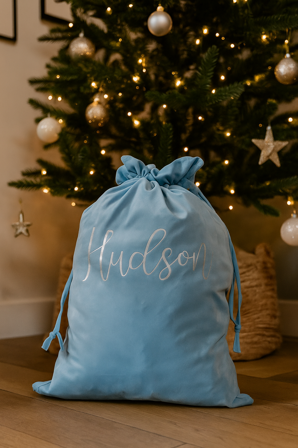 A light blue velvet Christmas sack with the name “Hudson” written in silver cursive sits on a wooden floor in front of a decorated Christmas tree adorned with gold and white ornaments and warm fairy lights.