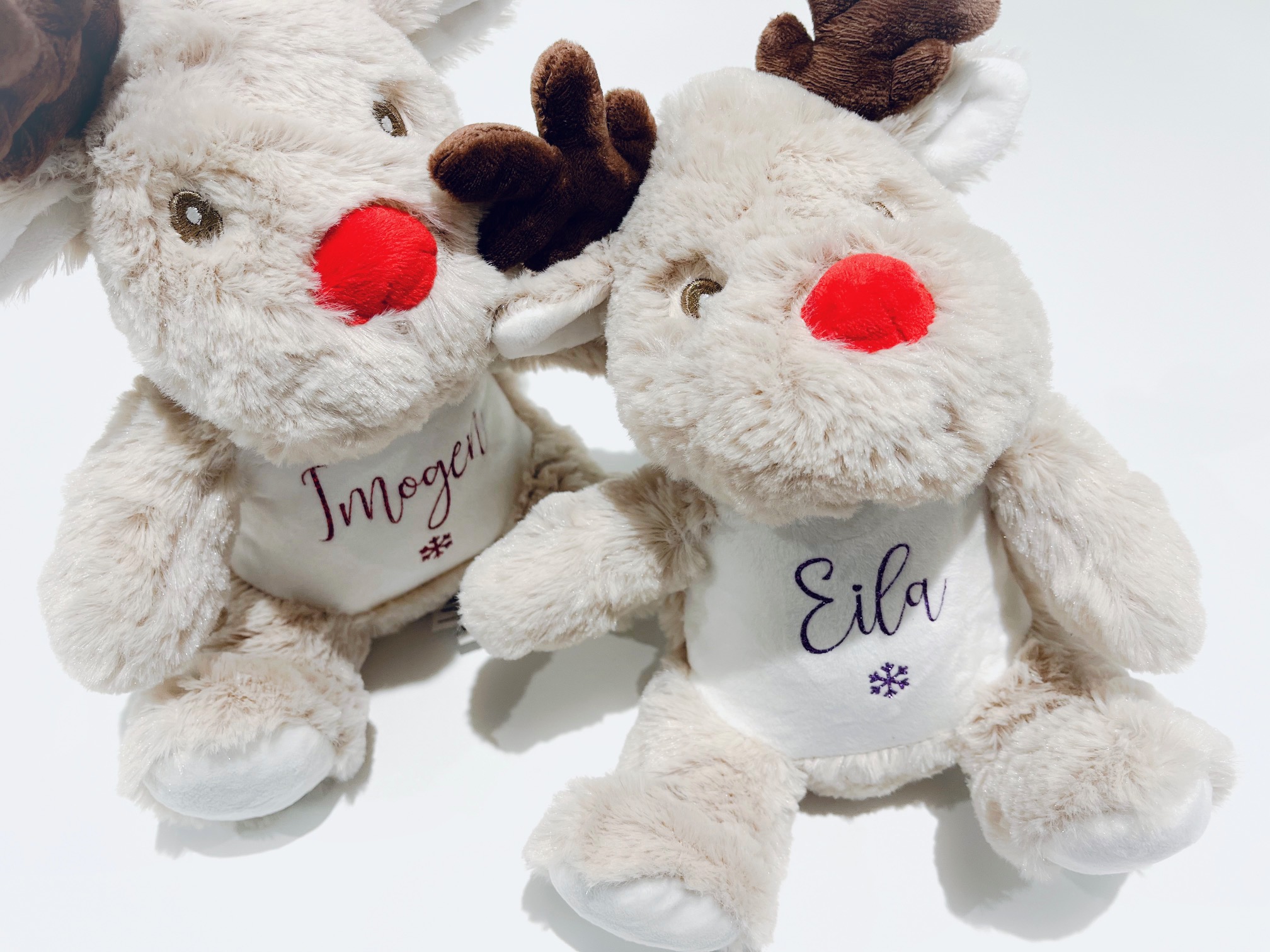 Two personalised soft plush reindeer Christmas toys, sitting side by side on a white background. Each reindeer has a bright red nose, brown antlers, and a white patch on the chest with a custom personalised glitter name (Imogen and Eila) and a snowflake design. Perfect for personalised Christmas gifts and holiday keepsakes.