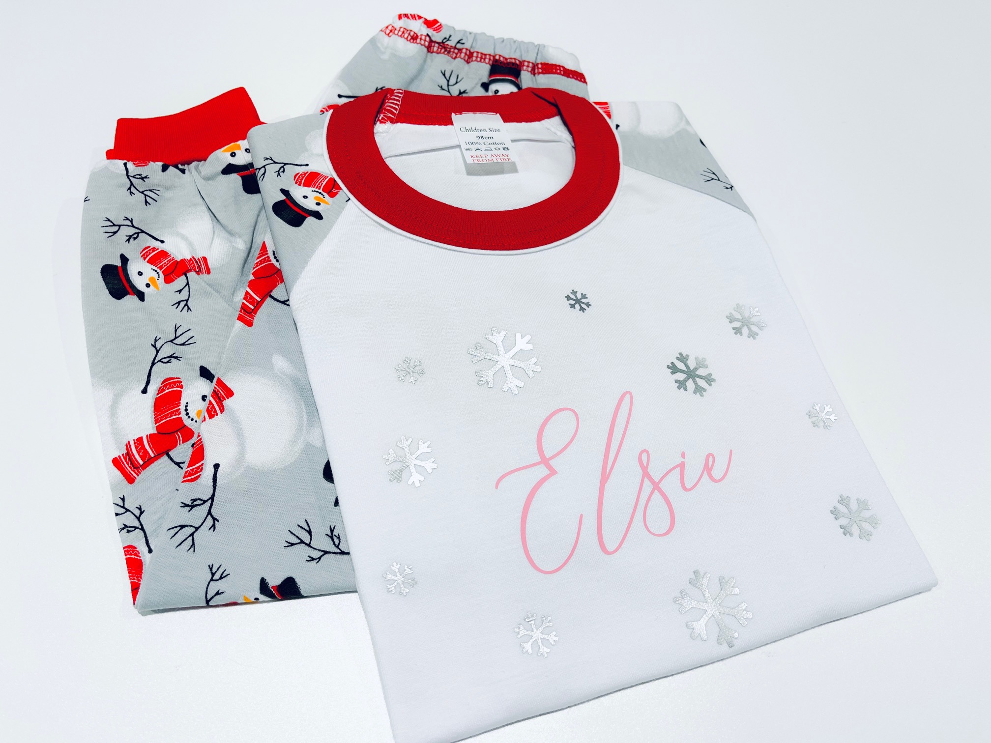 A set of children's Christmas pyjamas with a white raglan style top featuring a red collar and a printed snowflake design. The top is personalised with the name "Elsie" in a pink script font. The accompanying pyjama bottoms are patterned with repeating snowmen wearing red scarves on a grey background.