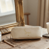 personalised cream accessory case with the name "Sue" written in gold script is resting on a rustic wooden vanity. Next to it is a steaming cup of tea, a silver tube of lotion or cream, and a delicate silver necklace. To the left is a vintage, ornate gold-framed mirror reflecting a window, and a glass jar containing cotton rounds. A makeup brush lies nearby. The overall setting is cosy and softly lit, suggesting a relaxing morning routine.