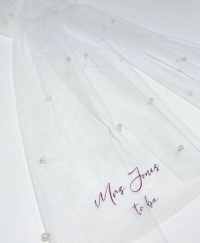 A short, two-tier white tulle bridal veil featuring a large white satin bow at the top. The veil is embellished with scattered pearls and features the words 