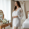 Bride in a white lace morning robe standing in a bright hotel suite with a wedding dress hanging in the background and white rose bouquet on a vanity