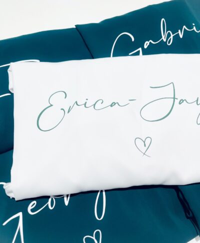 A stack of personalised forest green bridesmaid robes with white script personalisation. The top item is a white robe featuring the name 