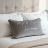 Grey personalised ‘Mr & Mrs Harris’ decorative cushion with pom-pom trim displayed on a neatly made bed with white pillows and neutral bedroom decor.
